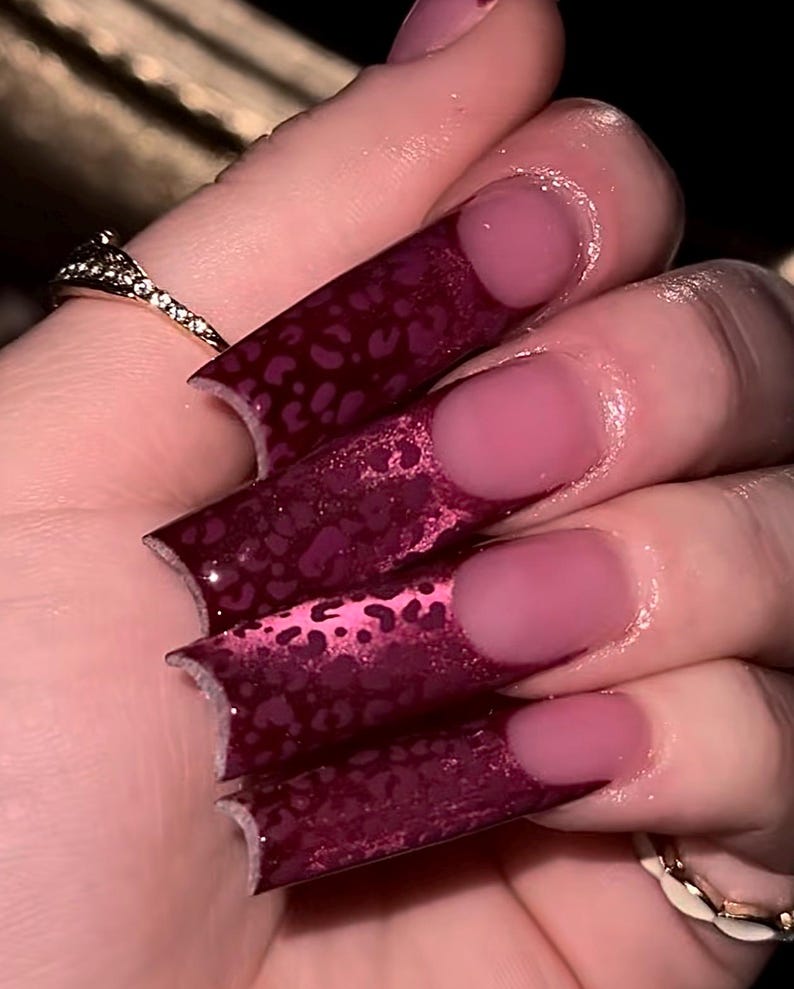 Pu&ograve; includere: Primo piano di unghie lunghe, quadrate, bordeaux con un motivo leopardato sulle punte. Le unghie hanno una finitura lucida e una base rosa. &Egrave; visibile anche un anello d'oro con un motivo a diamante.