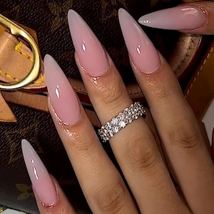 May include: Close-up of long, almond-shaped nails with a pink and white ombre design. The nails are adorned with a thin, gold glitter outline. A silver ring with multiple small stones is on the ring finger.