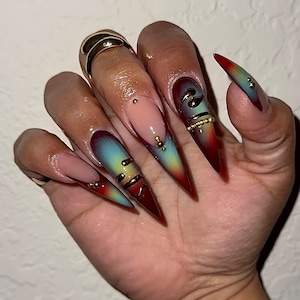 May include: Long, stiletto-shaped artificial nails with a gradient design. The nails transition from red to teal and yellow, adorned with gold accents and small jewels. Some nails feature gold spiral designs and a nude base.