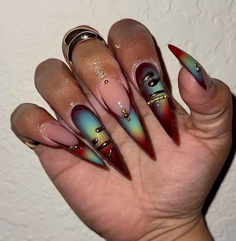 May include: Long, stiletto-shaped artificial nails with a gradient design. The nails transition from red to teal and yellow, adorned with gold accents and small jewels. Some nails feature gold spiral designs and a nude base.