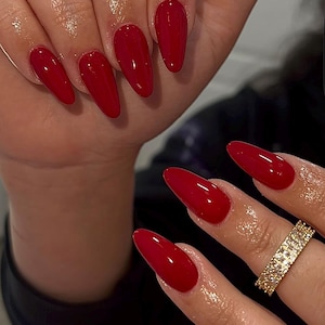 May include: Close-up of hands with long, glossy, red nail polish. The nails are oval-shaped. One hand has a gold ring with small, square-cut stones. The skin appears moisturized.