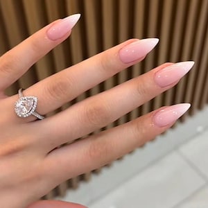 May include: A hand with long, pointed nails featuring a pink and white ombre design. A silver ring with a pear-shaped diamond is on the ring finger. The nails have a glossy finish.