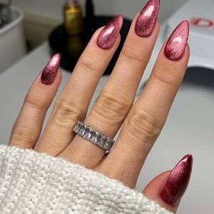 May include: A silver-toned eternity band ring featuring a row of clear, rectangular baguette-cut stones. The ring is worn on a finger with long, almond-shaped nails finished in a shimmering, metallic dark pink cat-eye polish.