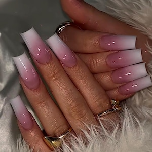 Puede incluir: Primer plano de una mano con uñas acrílicas largas y cuadradas. Las uñas presentan un diseño degradado, pasando de una punta blanca a una base rosa suave. Anillos dorados adornan los dedos. Las uñas parecen brillantes y bien cuidadas.
