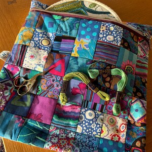 May include: A patchwork fabric zipped pouch with a round embroidery hoop, scissors, and green embroidery floss. The pouch is made of colourful fabric squares with floral and geometric patterns.