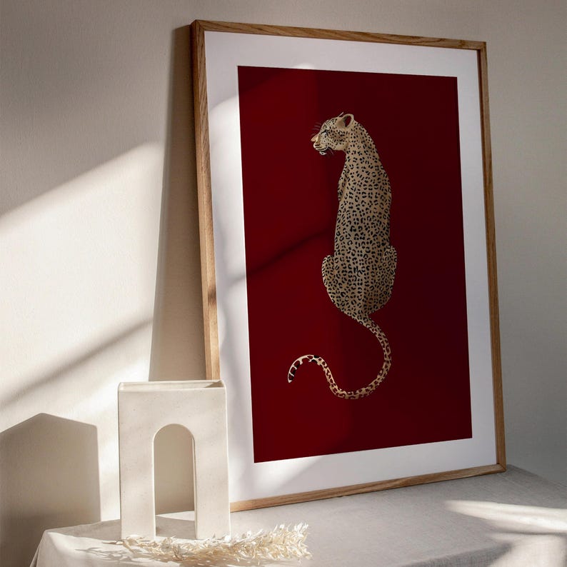 May include: A framed print of a leopard sitting on a red background. The leopard is facing to the left and has a long tail.