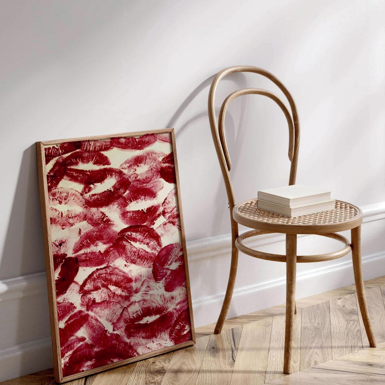 May include: A framed print of a red and white abstract design featuring multiple red lips printed on a white background. The print is leaning against a wooden chair with a woven seat.