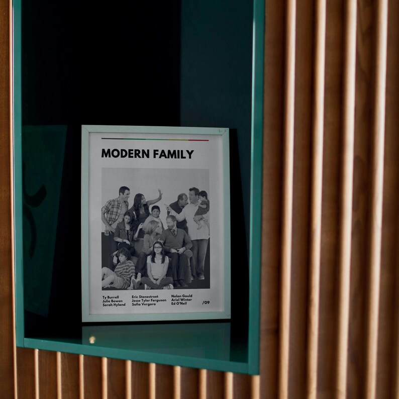 May include: A framed poster with the text "MODERN FAMILY" at the top. The black and white image features a group of people. The poster is in a light-colored frame and is displayed in a teal-colored shelf.