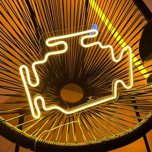 Pode incluir: Um sinal de néon amarelo em forma de luz de aviso do motor de um carro, em uma cadeira de vime preta. O sinal está aceso e projeta um brilho quente.