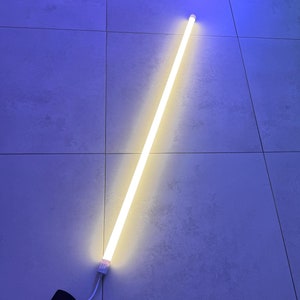 Puede incluir: Un tubo de luz LED blanco con un cable blanco y un enchufe negro. El tubo de luz está sobre una superficie de azulejos de color azul claro.