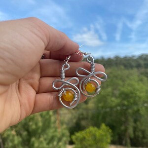 Puede incluir: Un par de pendientes de alambre plateado con cuentas de piedras preciosas amarillas. Los pendientes tienen forma de remolino.