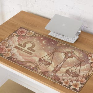 Könnte beinhalten: Eine rechteckige Schreibtischunterlage mit einem Waage-Tierkreiszeichen-Design. Die Matte hat ein rosa-beiges Farbschema mit floralen und himmlischen Elementen. Die Waage-Waage ist prominent dargestellt, zusammen mit Sternen, Monden und Rosen. Ein Laptop und eine Tastatur befinden sich auf dem Schreibtisch.
