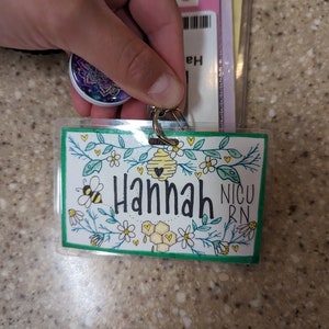 May include: A green and white ID badge with a floral design and a beehive. The badge says "Hannah NICU RN" in black lettering.