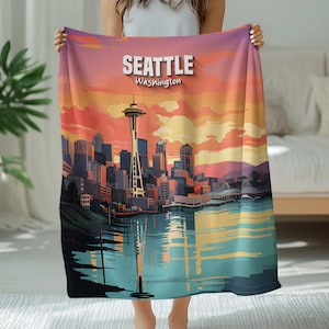 Könnte beinhalten: Eine Decke mit einer farbenfrohen Illustration von Seattle, Washington, mit der Space Needle und der Skyline, die sich im Wasser spiegelt. Die Decke hat ein Sonnenuntergangsfarbschema und den Städtenamen in Weiß.