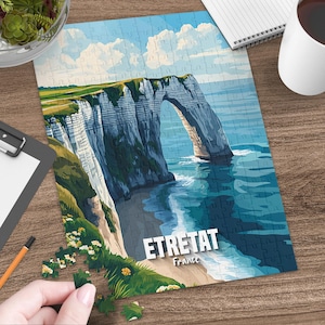 Könnte beinhalten: Ein fertiges Puzzle mit einer malerischen Ansicht von Étretat, Frankreich, mit einem natürlichen Bogen in den Klippen. Das Puzzle zeigt einen blauen Himmel mit weißen Wolken, grünes Gras und türkisfarbenes Wasser. Der Text "ETRETAT France" ist zu sehen.