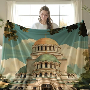May include: A large, rectangular blanket featuring a detailed illustration of a cathedral with gold domes and green roofs. The blanket has a blue sky background with clouds and is trimmed with tree branches.