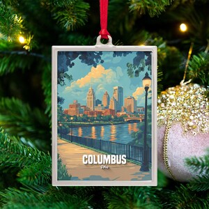 May include: A rectangular Christmas ornament featuring a scenic view of Columbus, Ohio, with a river, buildings, and a lamppost. The ornament has a silver frame and a red ribbon for hanging. The word "Columbus" is printed on the bottom.