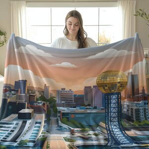 May include: A soft, colorful blanket featuring a cityscape design with a golden sphere structure. The blanket showcases a skyline with buildings, clouds, and a sunset color scheme. The blanket is held up by a person.