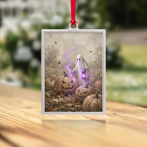 May include: A rectangular silver-framed ornament featuring a ghost holding a purple lantern, standing among carved jack-o'-lanterns and fallen autumn leaves. The ghost emits a purple mist, and the ornament hangs from a red ribbon.
