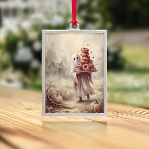 May include: A rectangular silver-framed ornament hanging from a red ribbon. The print depicts a ghost in a white sheet carrying a stack of glazed donuts through a field of pumpkins near a house, rendered in muted sepia and red tones.