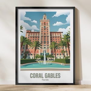 May include: A framed art print featuring a stylized illustration of the Biltmore Hotel in Coral Gables, Florida. The image depicts a tall pink building with a central tower, palm trees, and a fountain in the foreground under a blue sky.