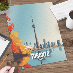 Può includere: Un puzzle completo raffigurante lo skyline di Toronto, con la CN Tower, edifici e lungomare. Il puzzle ha uno sfondo di cielo blu, con alberi autunnali arancioni e gialli. Il testo "Toronto Canada" è visibile.