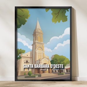 May include: Framed art print depicting the Santa Barbara D'Oeste church in Brazil. The illustration shows a beige church with a clock tower against a blue sky with clouds. The print is framed in black and rests on a wooden surface.