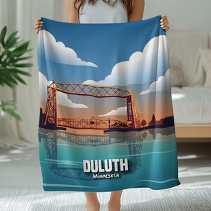 May include: A blue and white blanket featuring a scenic illustration of the Duluth, Minnesota, Aerial Lift Bridge reflected in the water. The blanket has a blue sky with white clouds and the text "Duluth Minnesota".