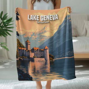 Könnte beinhalten: Eine Decke mit einer Landschaftsillustration des Genfer Sees, Schweiz. Das Design zeigt ein Schloss am See, Berge und den Text "Lake Geneva Switzerland" in Weiß. Die Farbpalette umfasst Blau-, Orange- und Weißtöne.