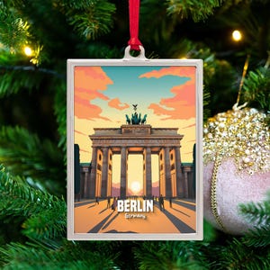 May include: A rectangular Christmas ornament featuring the Brandenburg Gate in Berlin, Germany, with a sunset sky. The ornament has a silver frame and a red ribbon for hanging. The word "BERLIN" is printed on the bottom.
