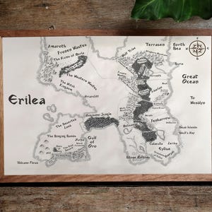 May include: A black and white hand-drawn map of the fictional land of Erilea. The map shows various regions, including the Frozen Wastes, the Western Wastes, the Gulf of Oro, and the Great Ocean. The map is framed in a brown wooden frame.