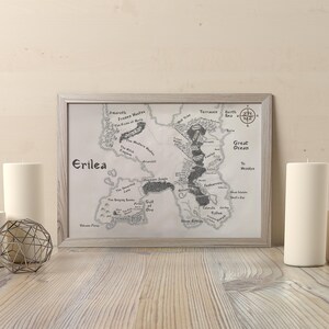 May include: A framed black and white map of the fictional land of Erilea. The map is surrounded by a white mat and a light gray frame. The map shows a large island with various rivers, lakes, and mountains. The text "Erilea" is printed in the top left corner of the map.