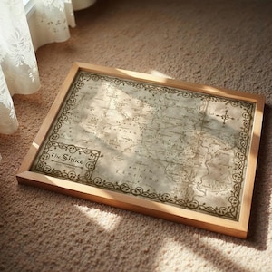 May include: A framed map of the Shire, featuring a vintage aesthetic with a light brown wooden frame. The map is detailed with place names and decorative borders, resting on a carpet.