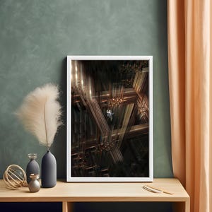 May include: A framed abstract art print with a white border. The artwork features a complex geometric design in shades of brown and gold, with a small figure suspended within. The print is displayed on a wooden surface, with decorative vases and a feather.