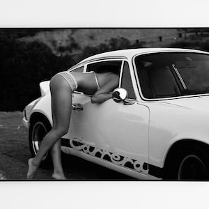 K&ouml;nnte beinhalten: Ein Schwarzwei&szlig;foto einer Frau, die sich in einen wei&szlig;en Porsche 911 Carrera lehnt. Das Auto steht auf einem Feldweg mit einem H&uuml;gel im Hintergrund.