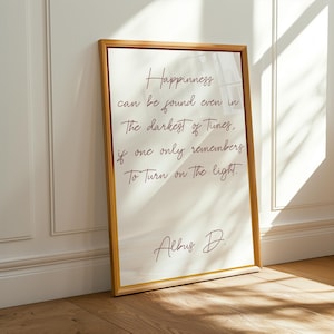 May include: A framed print with a gold-colored frame, leaning against a white wall. The print features handwritten text in brown ink that reads, "Happiness can be found even in the darkest of times, if one only remembers to turn on the light." Signed "Albus D."