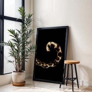 May include: A framed art print with a leopard tail design on a black background. The tail is rendered in brown and cream tones, offering a strong contrast. The artwork is positioned near a potted plant and a wooden stool.