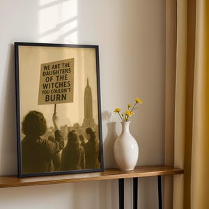 May include: Framed art print featuring a sepia-toned image of a protest. The print shows a group of people holding a sign with the text "WE ARE THE DAUGHTERS OF THE WITCHES YOU COULDN'T BURN" in front of a city skyline with the Empire State Building.