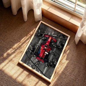 May include: A framed rectangular print featuring an overhead view of a bright red Formula One race car positioned in a pit stop area. The surrounding pit crew members are dressed in dark uniforms, and the ground is marked with white lines.