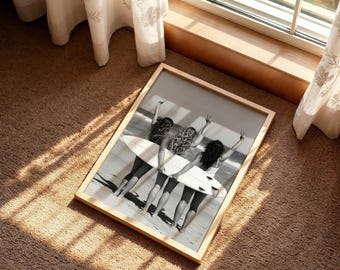May include: A rectangular framed wall art print featuring a black and white photograph of three individuals standing on a beach, holding a single surfboard horizontally. The individuals are seen from behind, with their arms raised in peace signs.