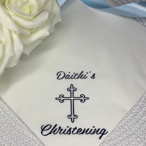 May include: White napkin with blue ribbon and the text "D&aacute;ith&iacute;'s Christening" in black lettering. A blue cross is centered above the text.