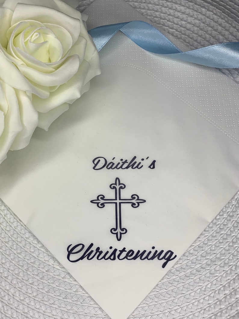 May include: White napkin with blue ribbon and the text "D&aacute;ith&iacute;'s Christening" in black lettering. A blue cross is centered above the text.