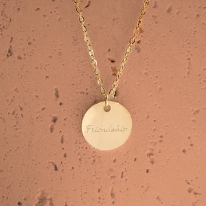 May include: A gold-toned necklace featuring a delicate chain and a circular pendant. The pendant has a smooth, polished surface with the word Friendship engraved in a cursive script font centered on the front of the round metal disc.