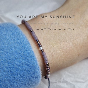 May include: A purple and white beaded bracelet with a black string tie. The bracelet has the words "YOU ARE MY SUNSHINE" written in braille.