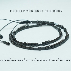 Op de afbeelding: Een zwarte kralen armband met een zilveren kraal accent. De armband is gemaakt van kleine, vierkante kralen en heeft een zwart koord met een knoop aan het einde. De tekst "I'D HELP YOU BURY THE BODY" staat boven de armband. De tekst ".--/..../.-../--/....-../.--/..../.-.." staat onder de armband.