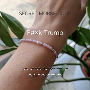Könnte beinhalten: Ein Perlenarmband mit hellrosa und durchscheinenden Perlen in einem Muster. Der Text SECRET MORSE CODE und F#xk Trump ist sichtbar, zusammen mit einer Morsecode-Sequenz, die durch Punkte und Striche auf dem Bild dargestellt ist.