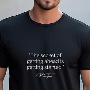 May include: Black t-shirt with the quote "The secret of getting ahead is getting started." in white text. The shirt also features the signature of Mark Twain. The shirt is worn by a person.