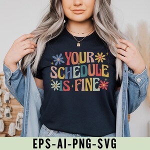 Pu&ograve; includere: T-shirt blu navy con la scritta "YOUR SCHEDULE IS FINE" in lettere colorate in stile retr&ograve;. Il design include accenti floreali in vari colori. La persona indossa anche una giacca di jeans. Il testo "EPS-AI-PNG-SVG" &egrave; visibile.