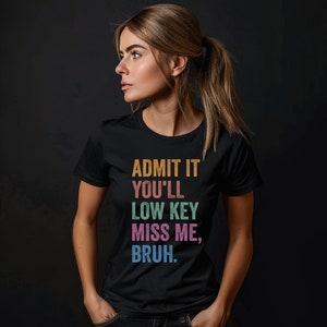 May include: Black crew neck t-shirt with the text "ADMIT IT YOU'LL LOW KEY MISS ME, BRUH" in orange, green, pink, and blue. The shirt is worn by a person with blonde hair tied back.