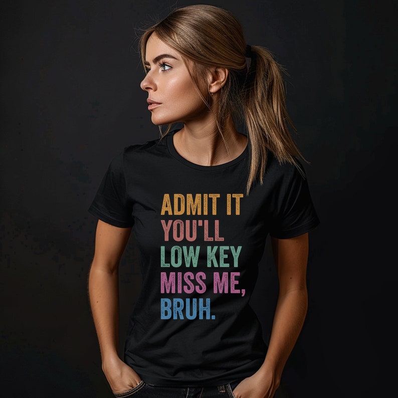 May include: Black crew neck t-shirt with the text "ADMIT IT YOU'LL LOW KEY MISS ME, BRUH" in orange, green, pink, and blue. The shirt is worn by a person with blonde hair tied back.